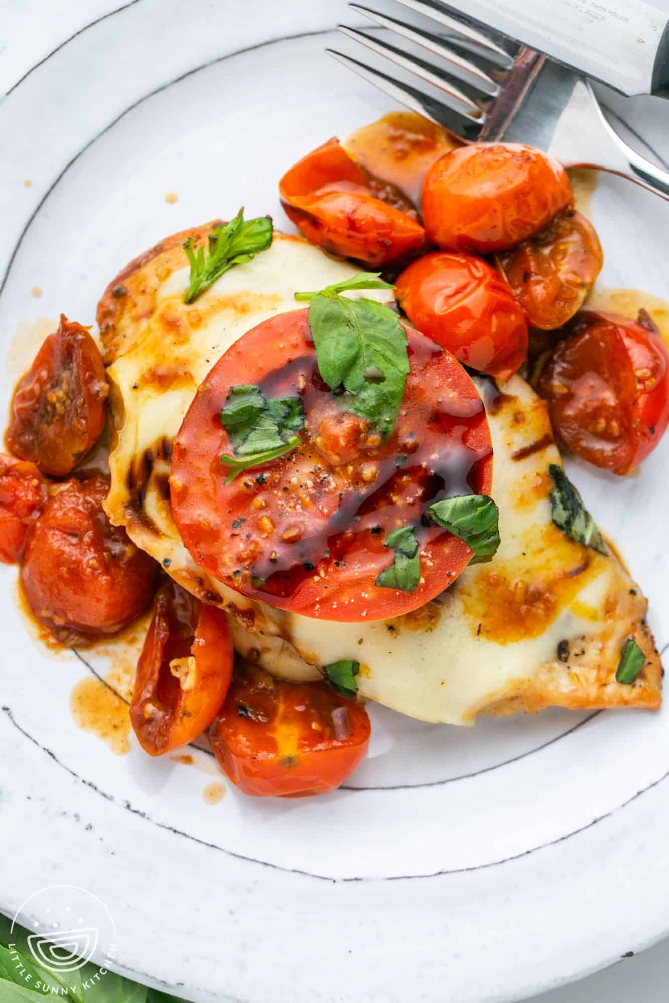 a white plate with a serving of caprese chicken with cherry tomatoes on it.