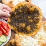 Overhead shot of zaatar and cheese manakish on a wooden board, with fresh cut tomato and cucumber on the side.