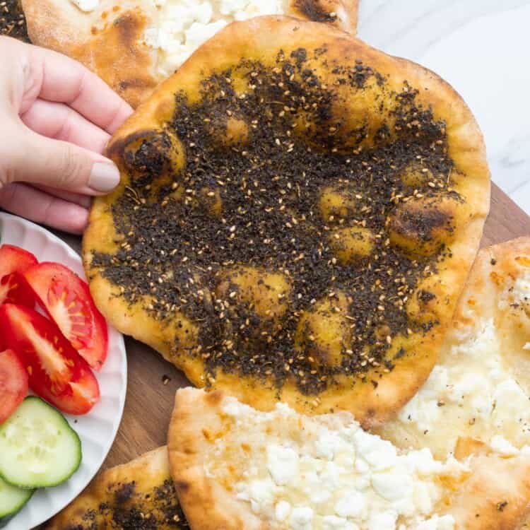 Overhead shot of zaatar and cheese manakish on a wooden board, with fresh cut tomato and cucumber on the side.