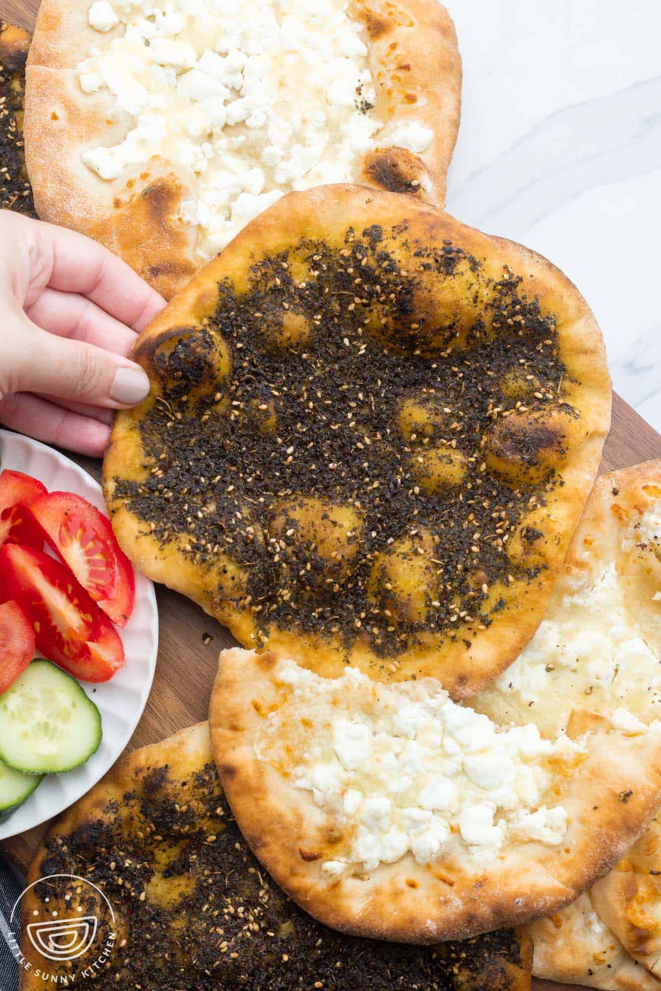 Overhead shot of zaatar and cheese manakish on a wooden board, with fresh cut tomato and cucumber on the side.