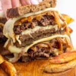 a patty melt, cut in half and stacked to show the inside. french fries are next to it on a wooden board. a hand is holding the sandwich.