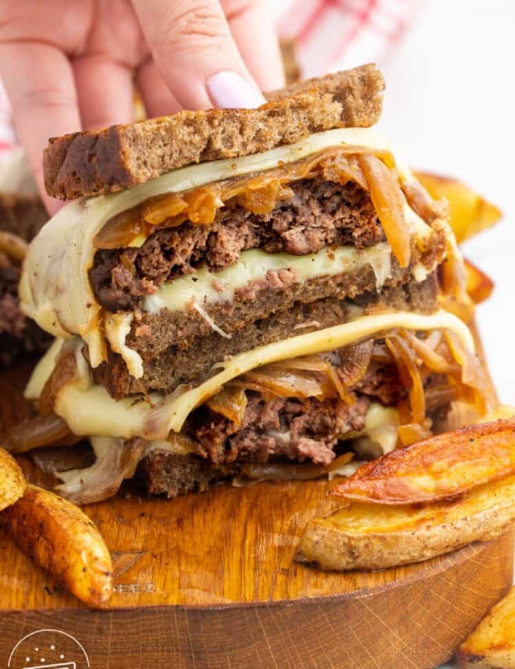 a patty melt, cut in half and stacked to show the inside. french fries are next to it on a wooden board. a hand is holding the sandwich.