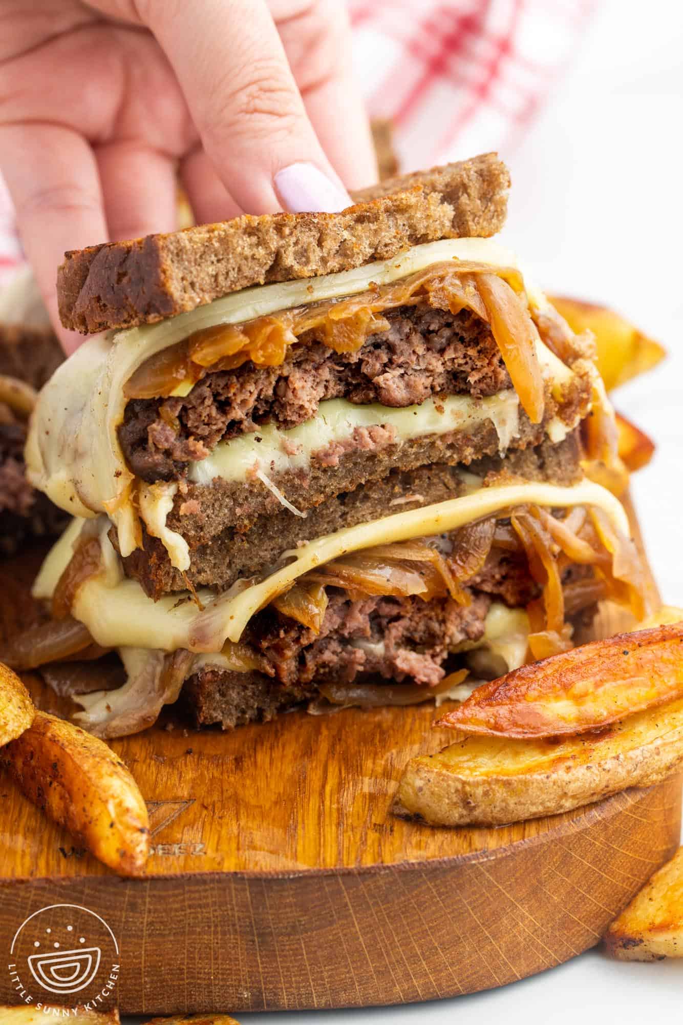 a patty melt, cut in half and stacked to show the inside. french fries are next to it on a wooden board. a  hand is holding the sandwich.