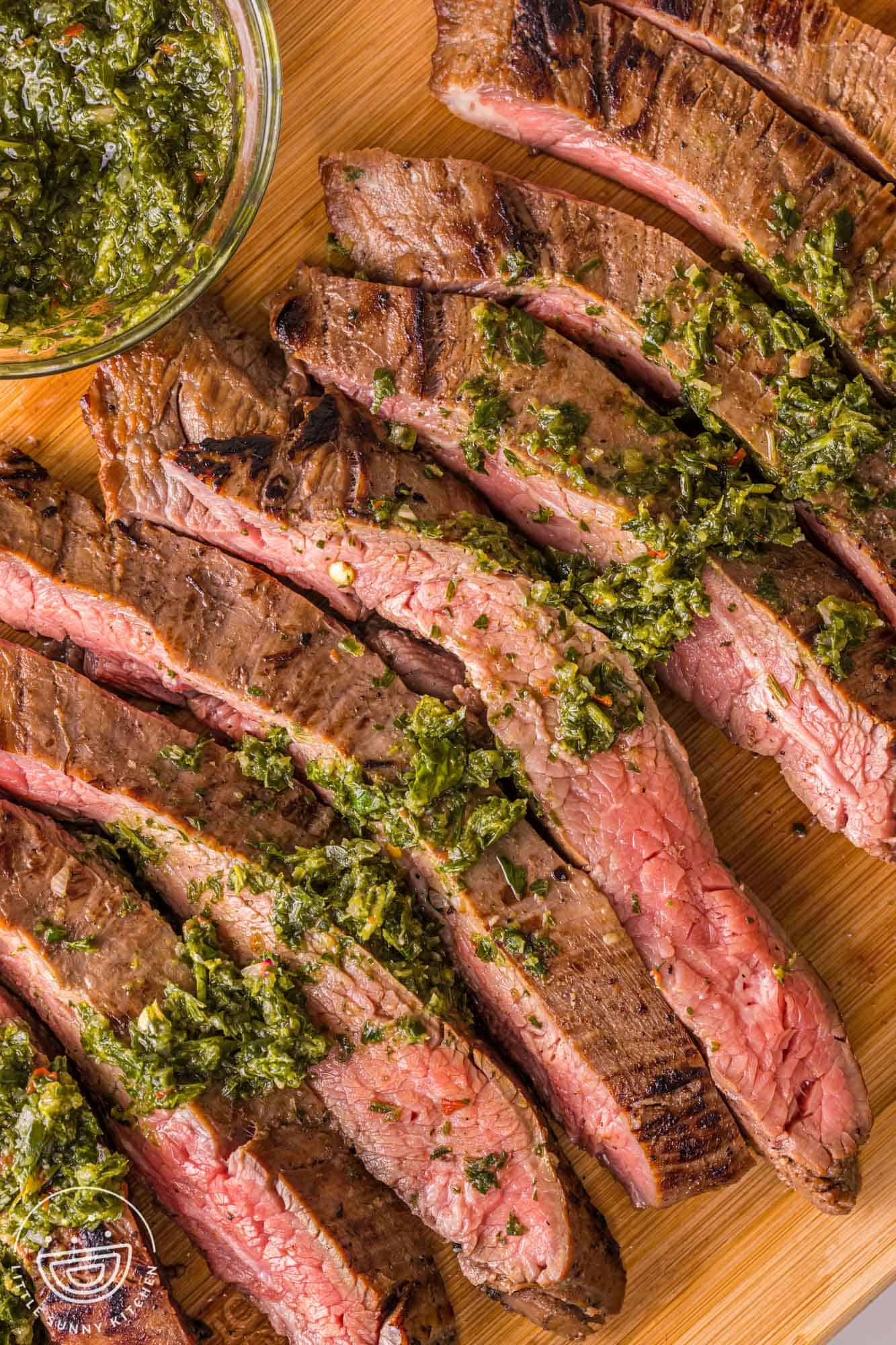 Overhead shot of sliced smoked flank steak, topped with fresh chimichurri sauce on a wooden cutting board
