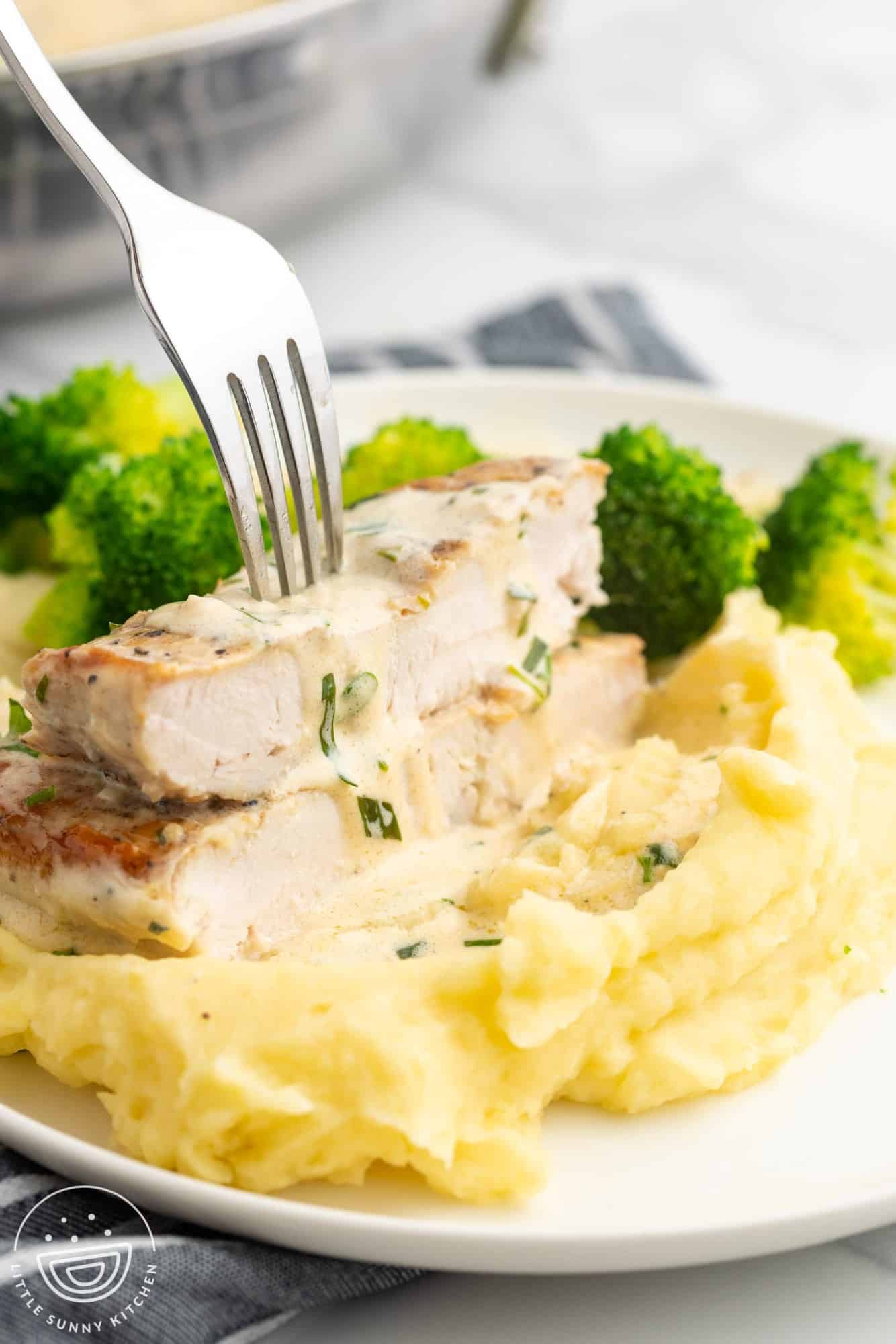 a fork holding sliced tarragon chicken on top of mashed potatoes, with broccoli in the background.