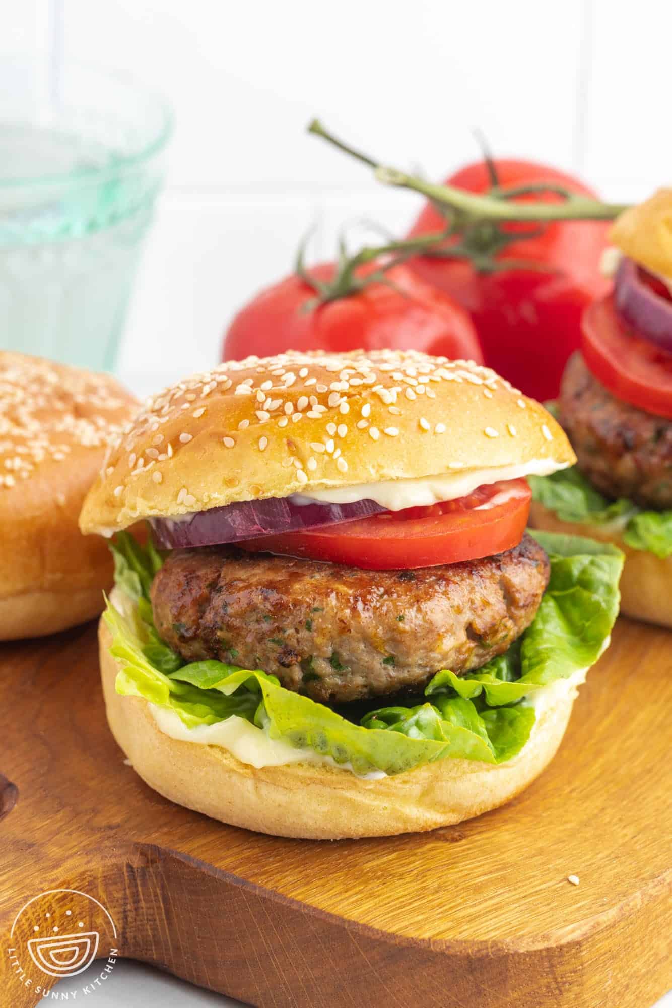 Turkey burger in a soft brioche bun, with lettuce, a slice of tomato, and red onion. Placed on a wooden cutting board with a bunch of tomatoes and brioche buns in the background.