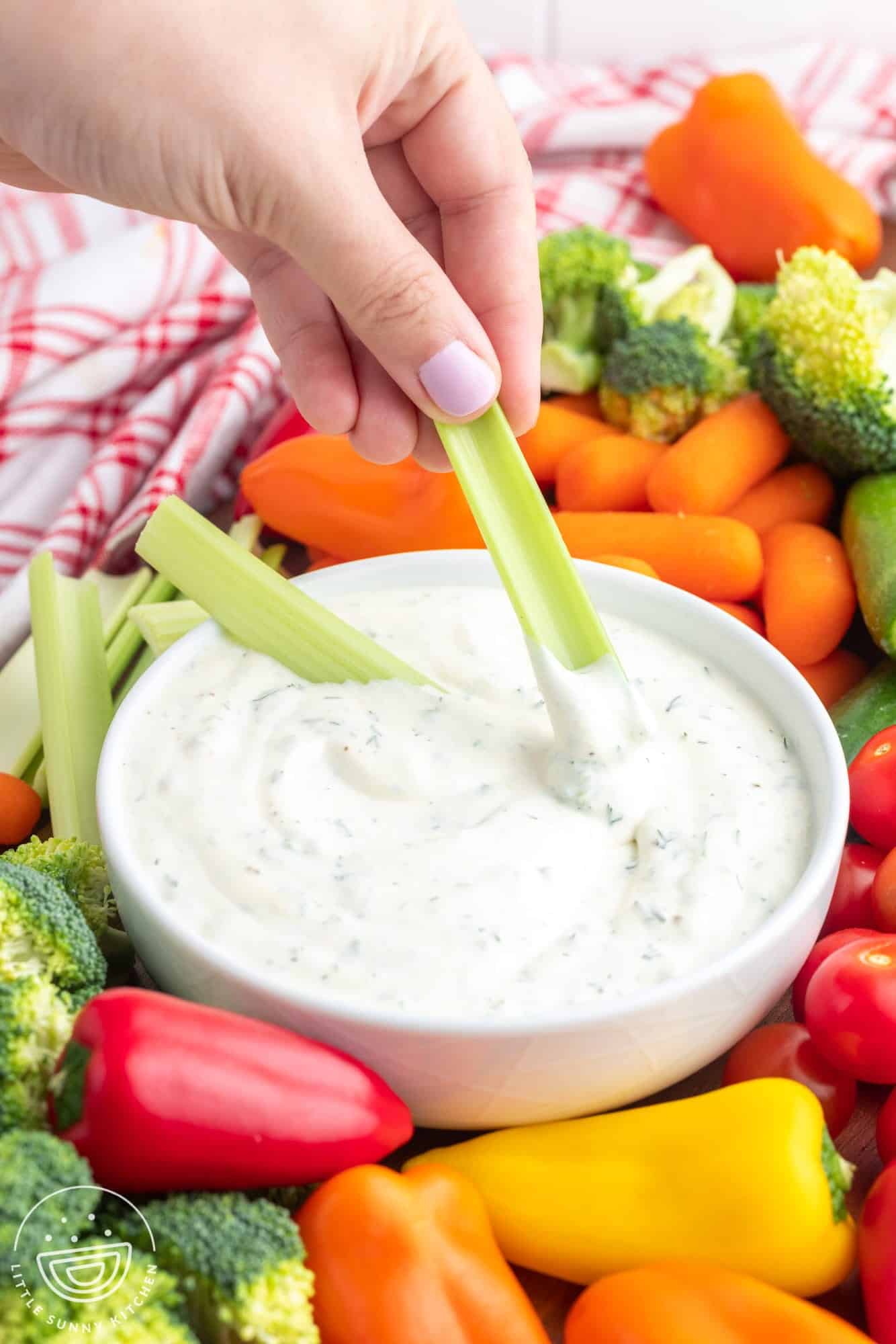 a hand dipping a celery stick into a bowl of easy veggie dip that is on a vegetable platter.