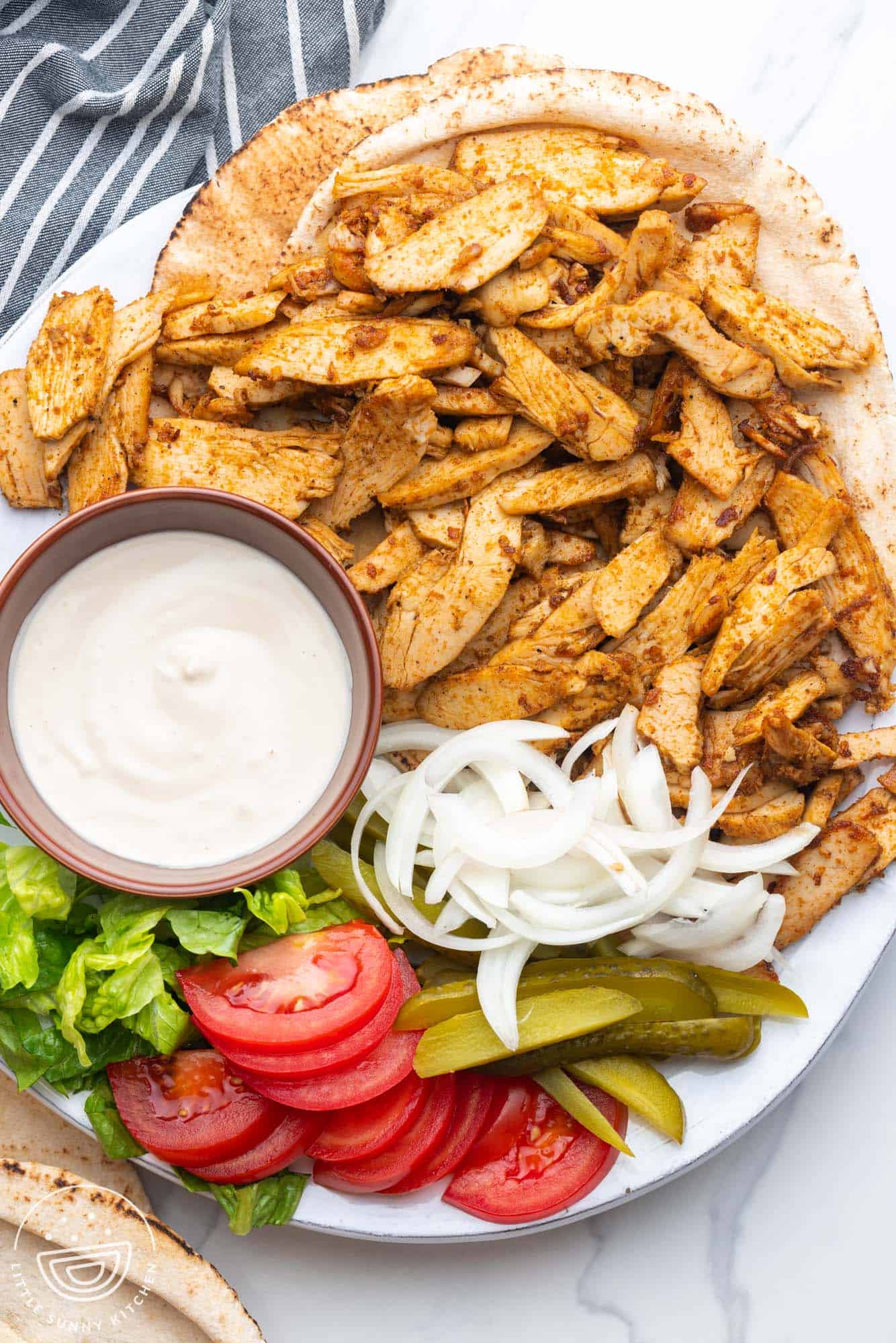 a platter of sliced chicken shawarma with creamy sauce, sliced tomatoes and onions, pickles, and shredded lettuce on the side.