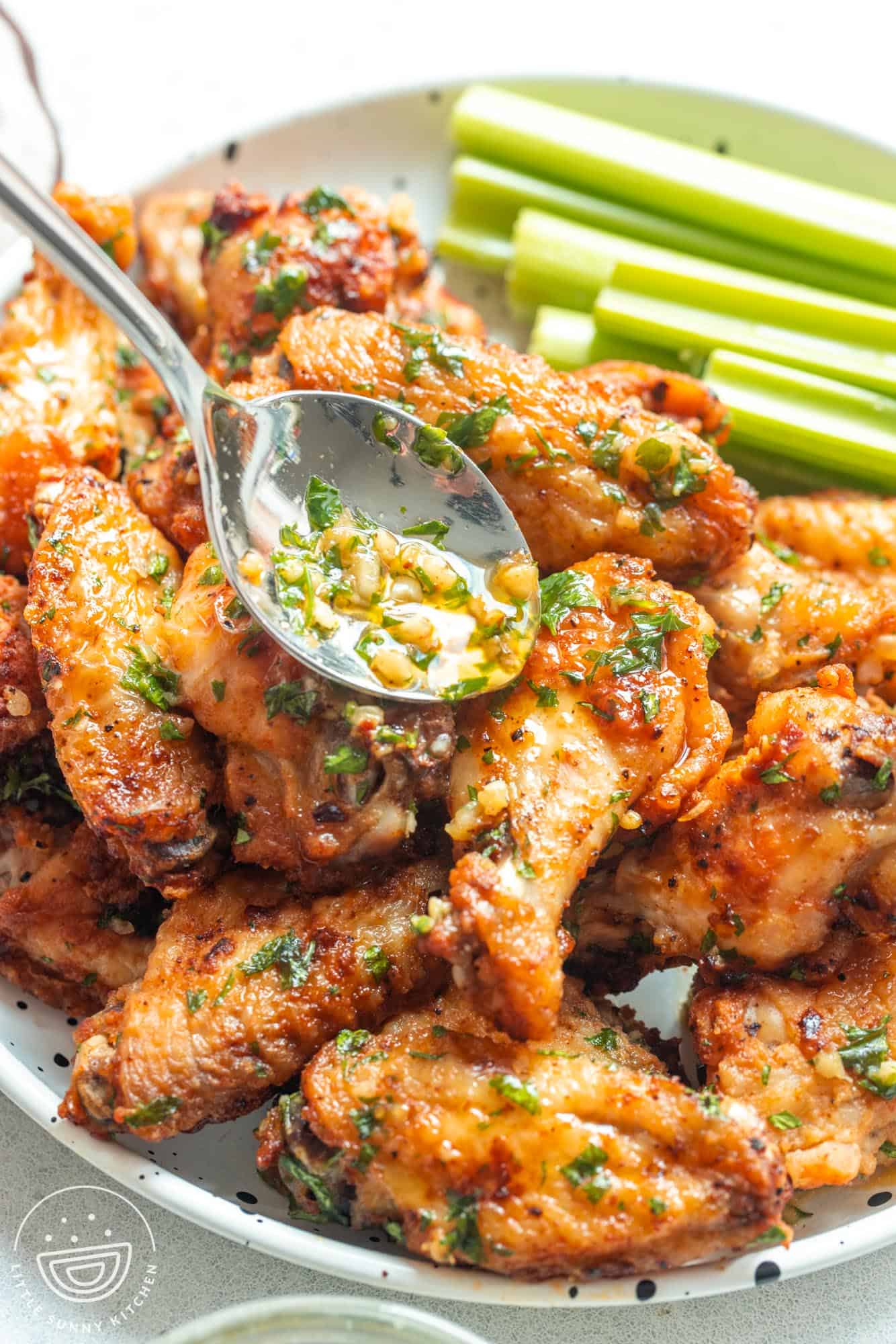 Drizzling garlic butter sauce over crispy wings in a plate, with celery sticks in the background