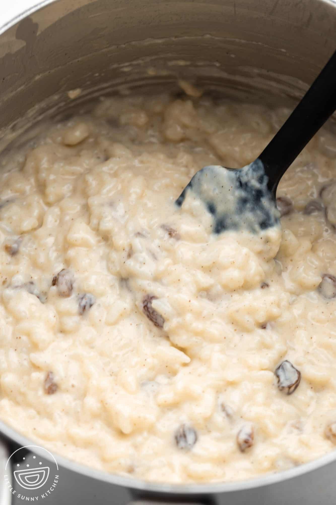 Rice pudding with raisins cooking in a saucepan, stirred with a spatula.