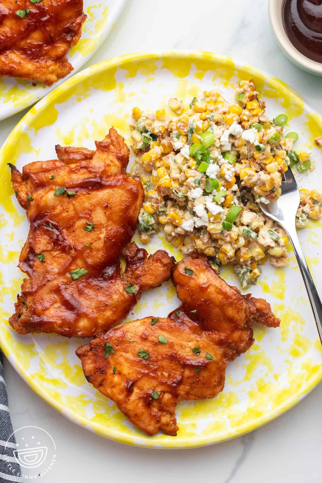 A dinner plate of two bbq chicken thighs and a side of corn salad.