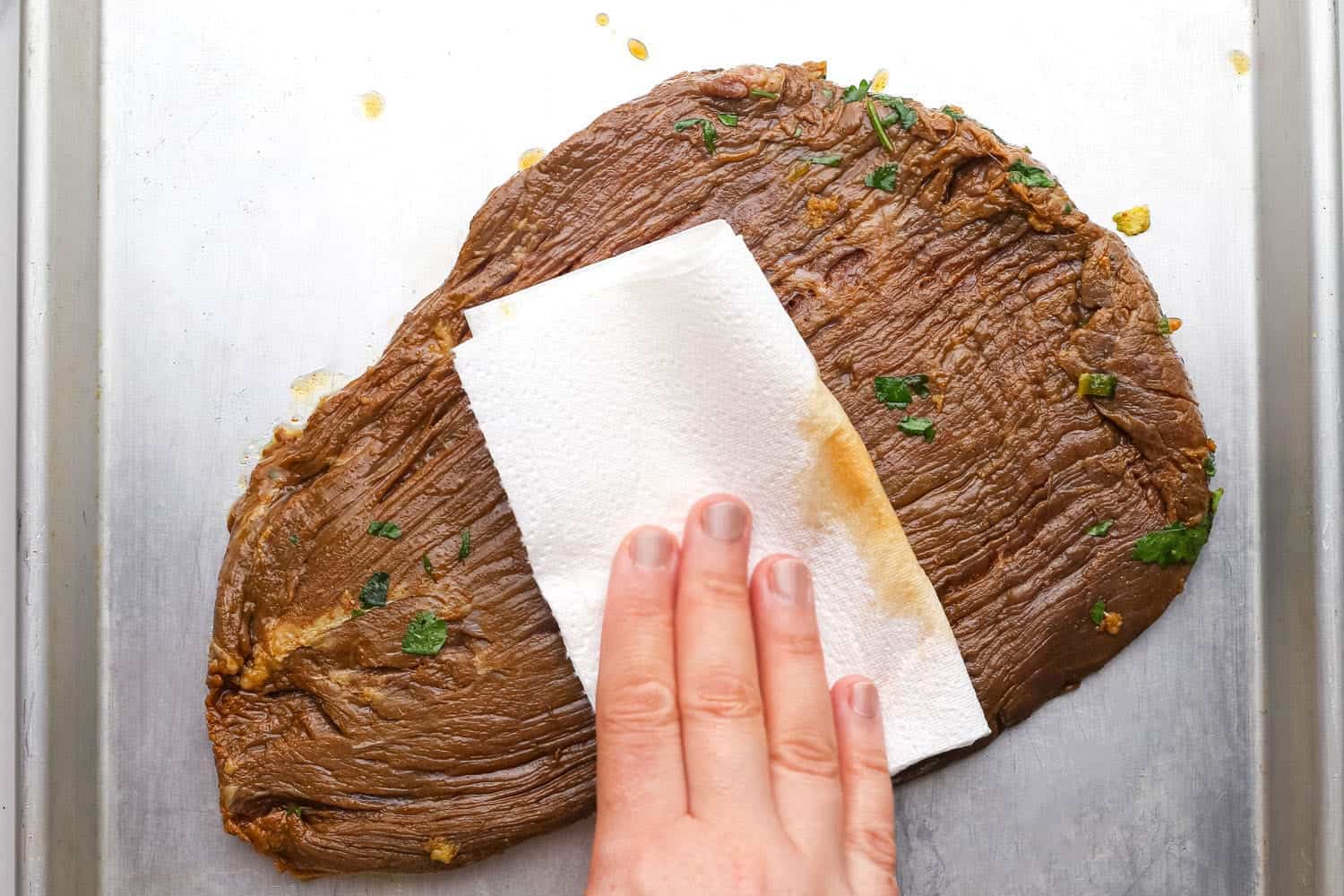 A marinated flank steak on a tray. a hand is drying the meat with a paper towel