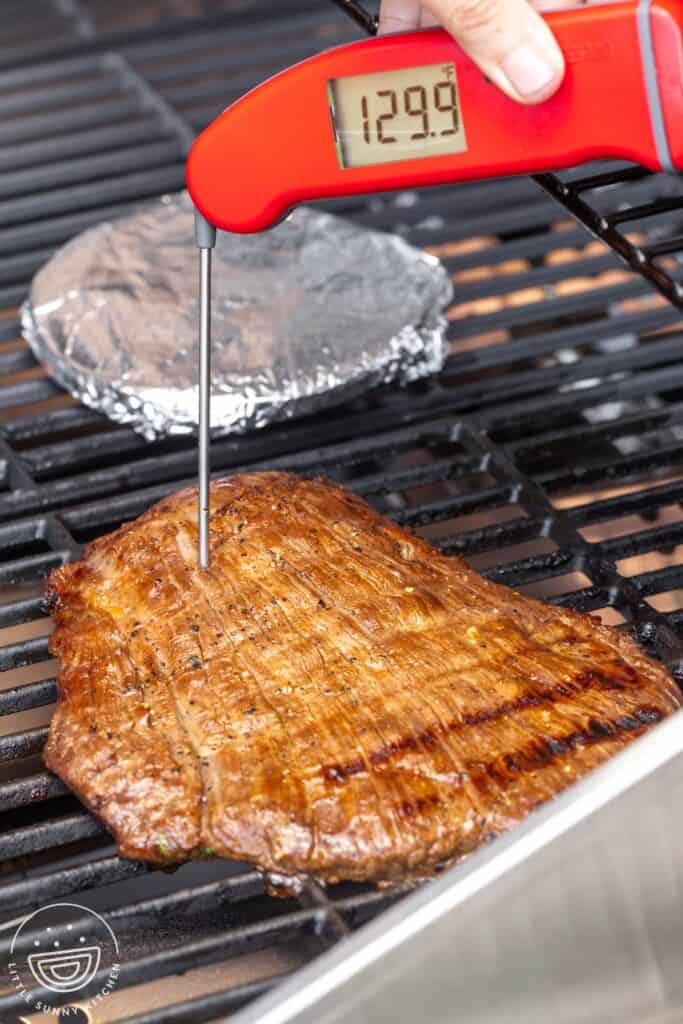 Carne asada being cooked on a grill. A thermometer is inserted, and reads 129.9°F