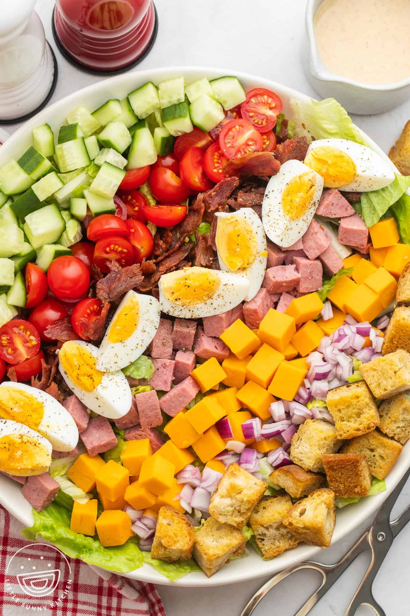 a large round bowl of salad topped with veggies, bacon, ham, eggs, cheese, in rows.
