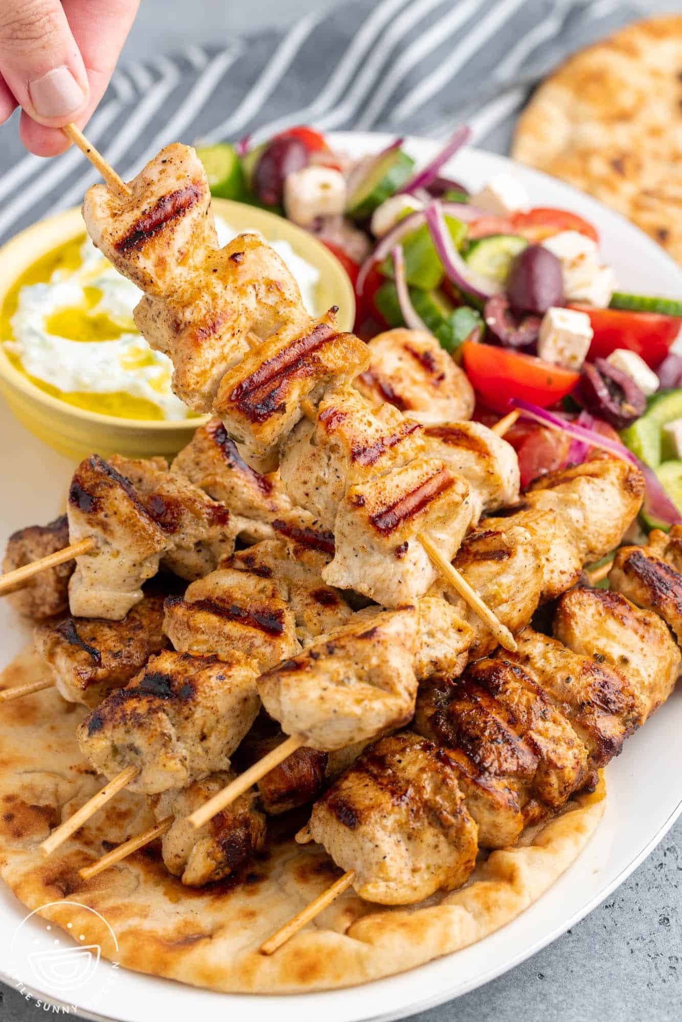 a chicken souvlaki platter. A hand is picking up a skewer of chicken from the plate.