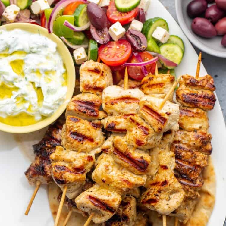 A platter of chicken souvlaki skewers on a pita, with a side of tzatziki sauce and greek salad.