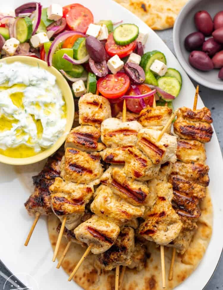 A platter of chicken souvlaki skewers on a pita, with a side of tzatziki sauce and greek salad.