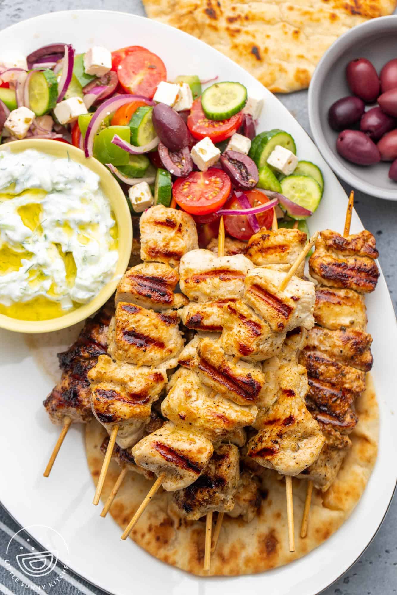 A platter of chicken souvlaki skewers on a pita, with a side of tzatziki sauce and greek salad.