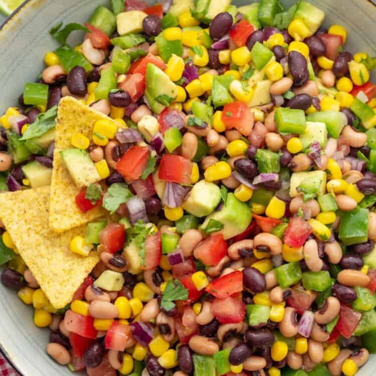 a serving bowl of cowboy caviar made with tomatoes, black eyed peas, peppers, corn, and avocado. There are two chips on the side of the bowl.