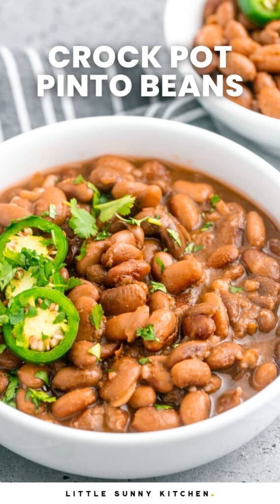 Pinto beans in a medium sized white bowl, topped with sliced jalapeno and cilantro. And overlay text that says "Crock pot pinto beans"