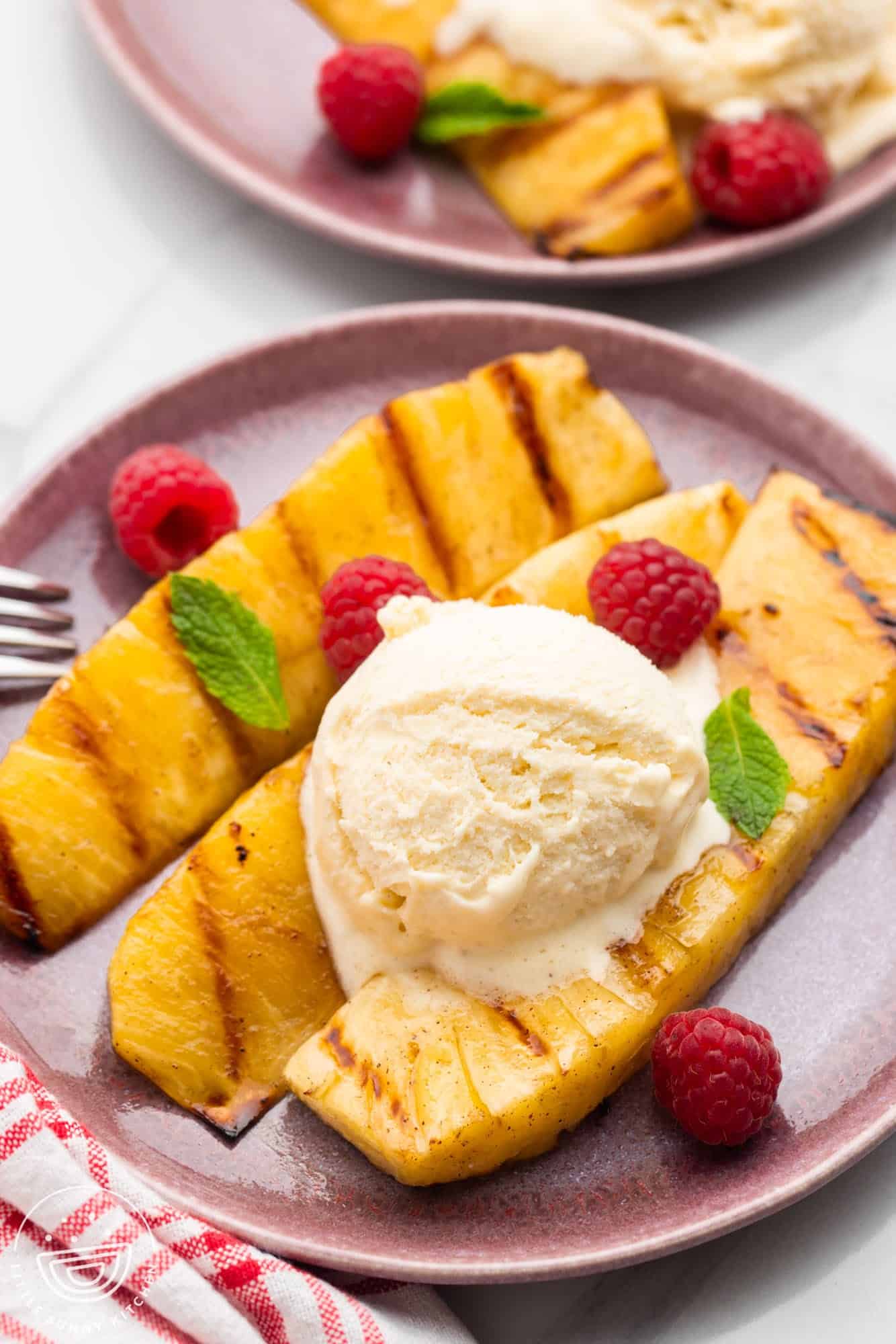 Grilled pineapple spears served on a pink plate with a scoop of ice cream and fresh raspberries and mint leaves