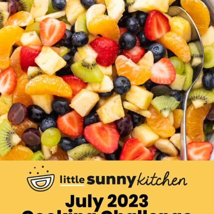 Overhead shot of a fruit salad with lime honey dressing in a large gray bowl, and a serving spoon. And overlay text that says "July 2023 Cooking Challenge"