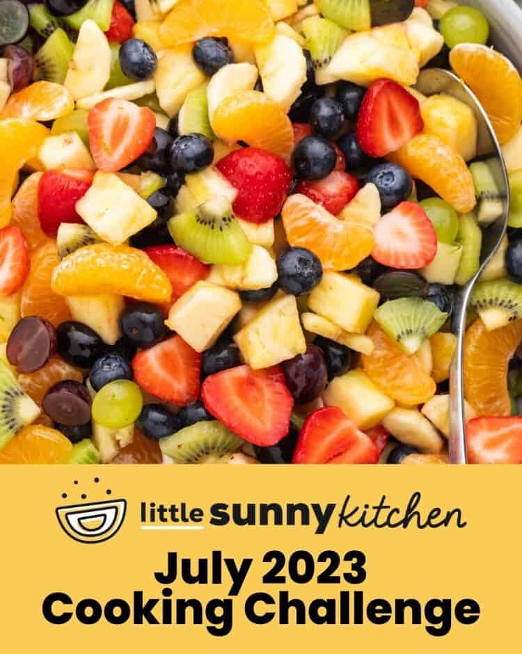 Overhead shot of a fruit salad with lime honey dressing in a large gray bowl, and a serving spoon. And overlay text that says "July 2023 Cooking Challenge"