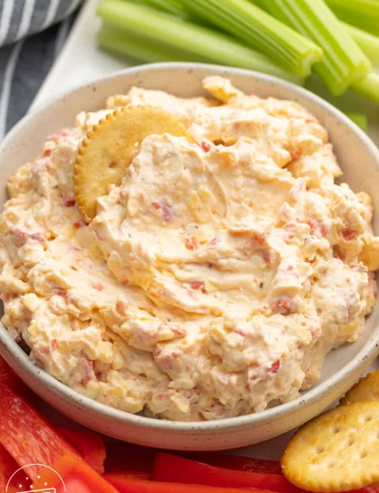 a bowl of pimento cheese next to celery sticks, red peppers, and crackers.