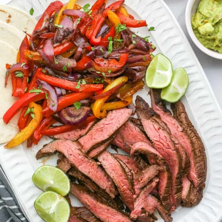 a large white platter of fajita veggies, sliced steak fajitas, and lime wedges. On the side are flour tortillas, sour cream, guacamole, and pico.