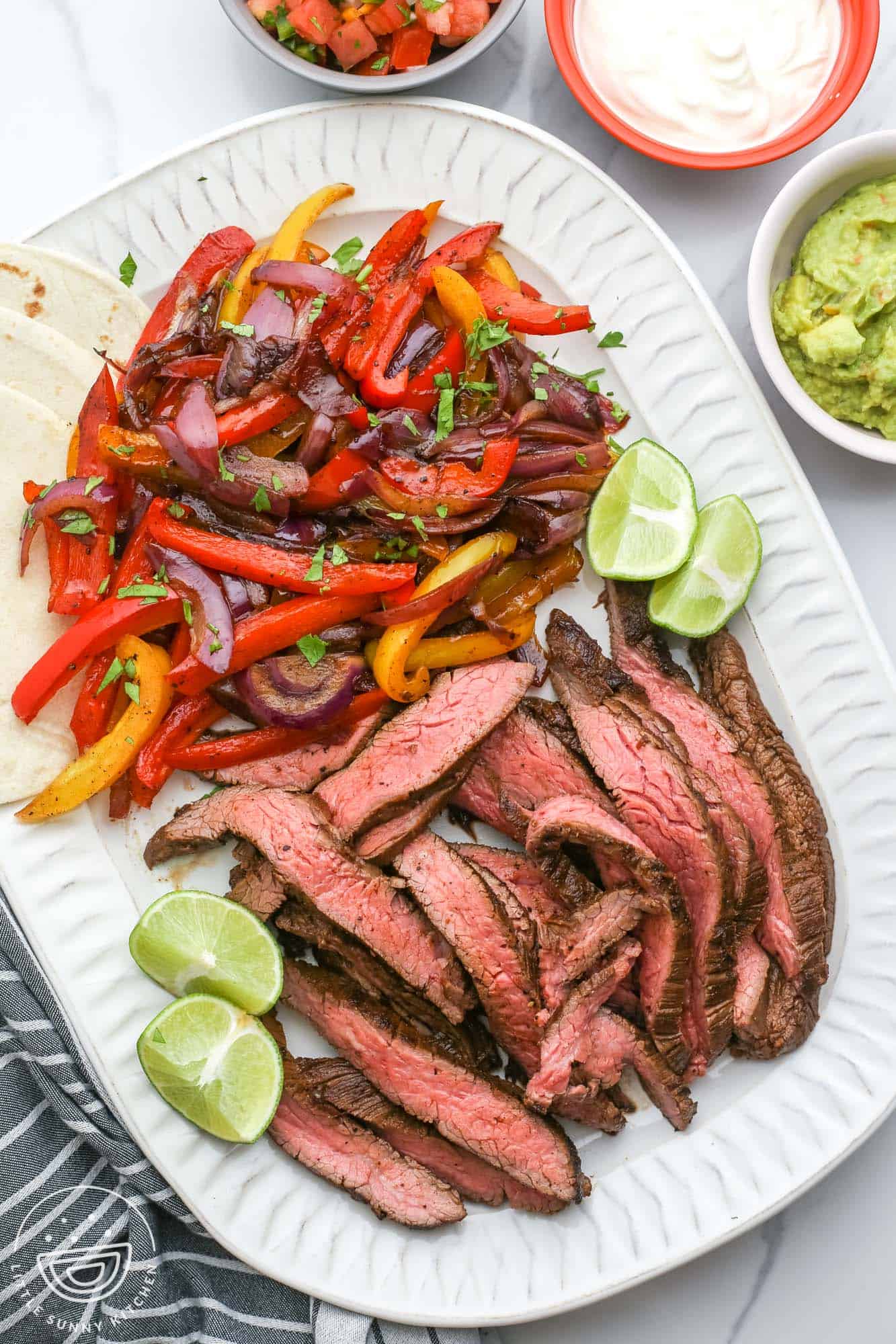 a large white platter of fajita veggies, sliced steak fajitas, and lime wedges. On the side are flour tortillas, sour cream, guacamole, and pico.