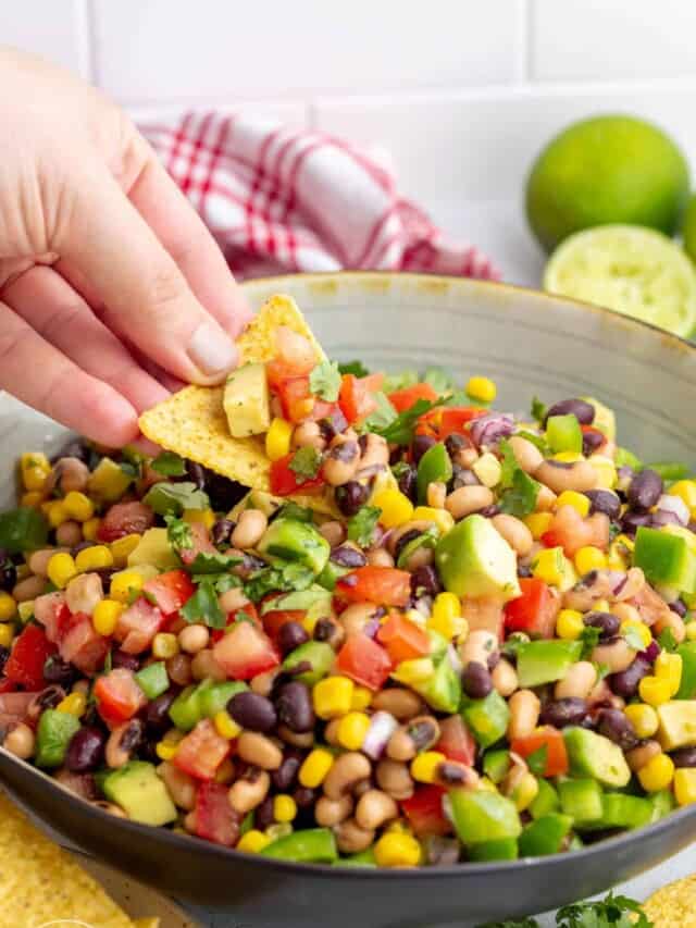 a hand holding a tortilla chip, dipping it into a large bowl of cowboy caviar.