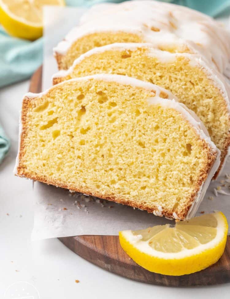 Close-up of three slices of lemon loaf cake with white glaze arranged on a wooden cutting board. A lemon wedge sits next to the cake, and crumbs are scattered on the parchment paper beneath it.