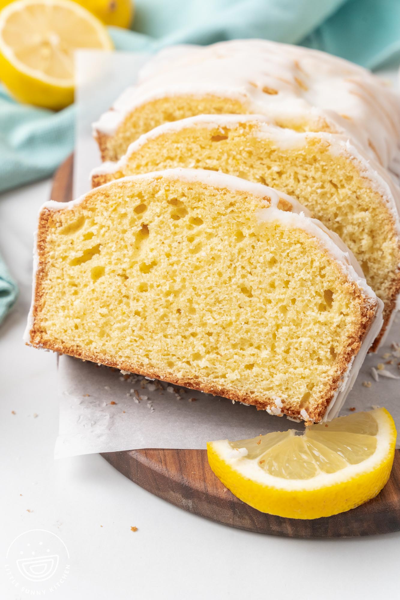 Close-up of three slices of lemon loaf cake with white glaze arranged on a wooden cutting board. A lemon wedge sits next to the cake, and crumbs are scattered on the parchment paper beneath it.