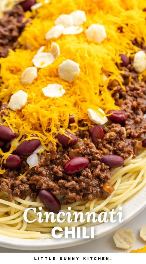 a plate of spaghetti topped with chili, beans, cheese, and oyster crackers. Text overlay says "cincinnati chili"