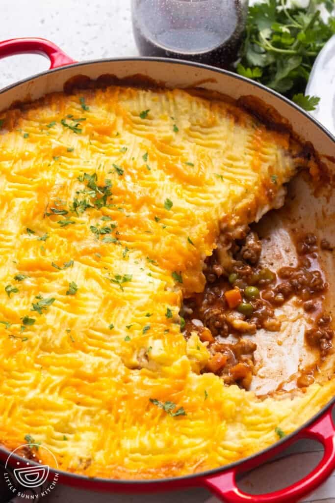 a red, round baking dish filled with cottage meat pie topped with cheesy mashed potatoes.