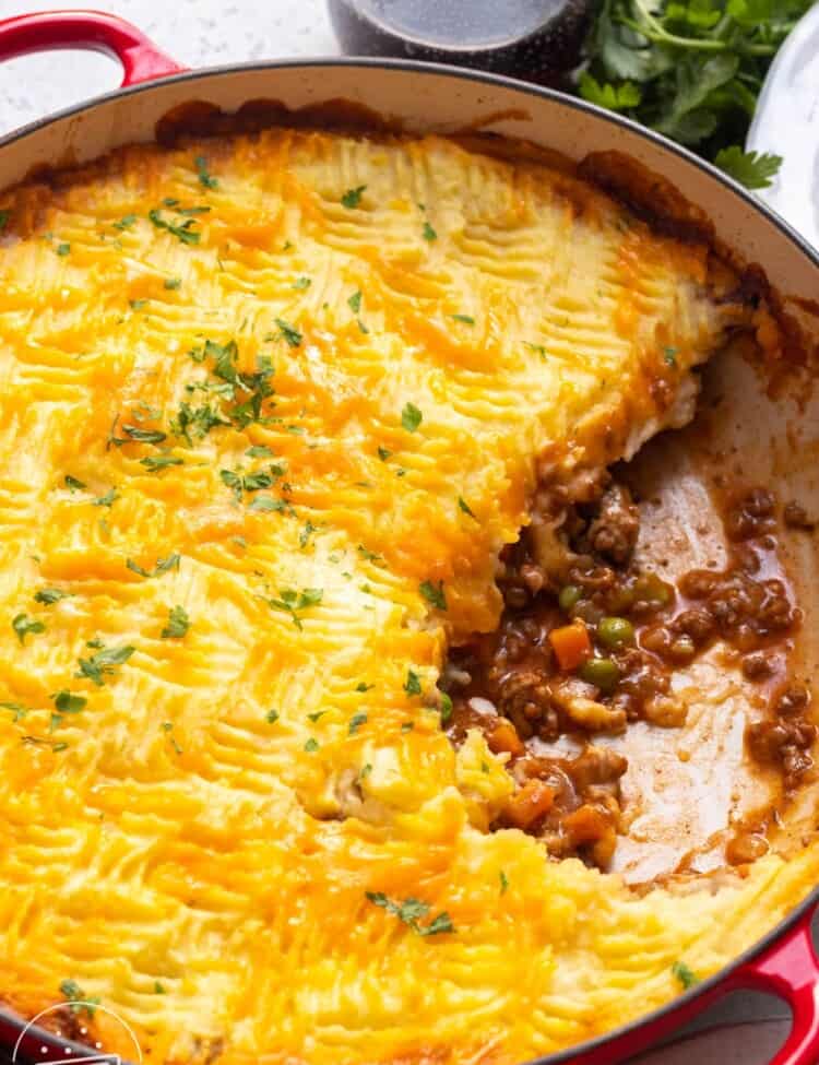 a red, round baking dish filled with cottage meat pie topped with cheesy mashed potatoes.