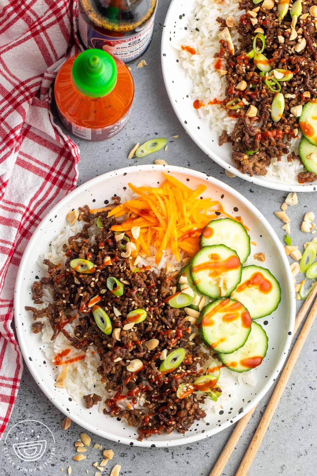 A plate of rice topped with korean seasoned ground beef, sliced cucumbers and shredded carrot, drizzled with sriracha