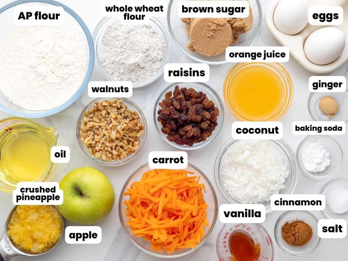 The ingredients needed to make morning glory muffins, including shredded carrot, coconut, walnuts, raisins, and pineapple, all in separate bowls, arranged on a counter.