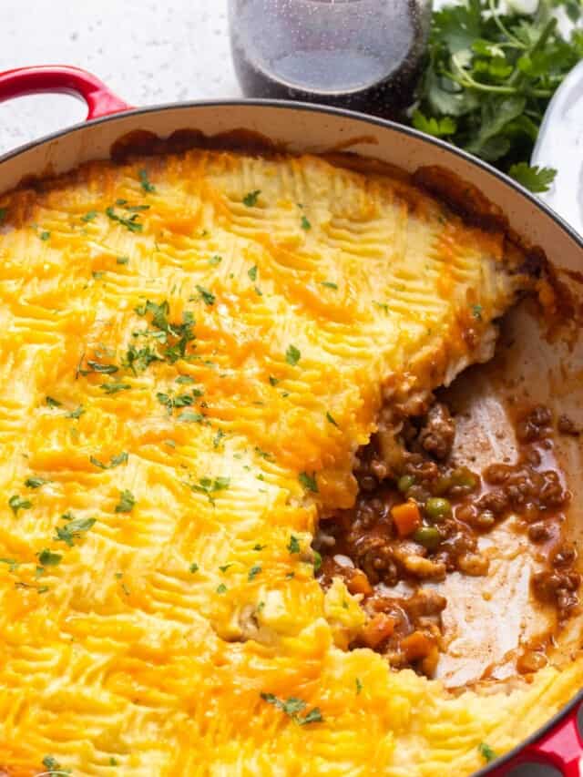a red, round baking dish filled with cottage meat pie topped with cheesy mashed potatoes.