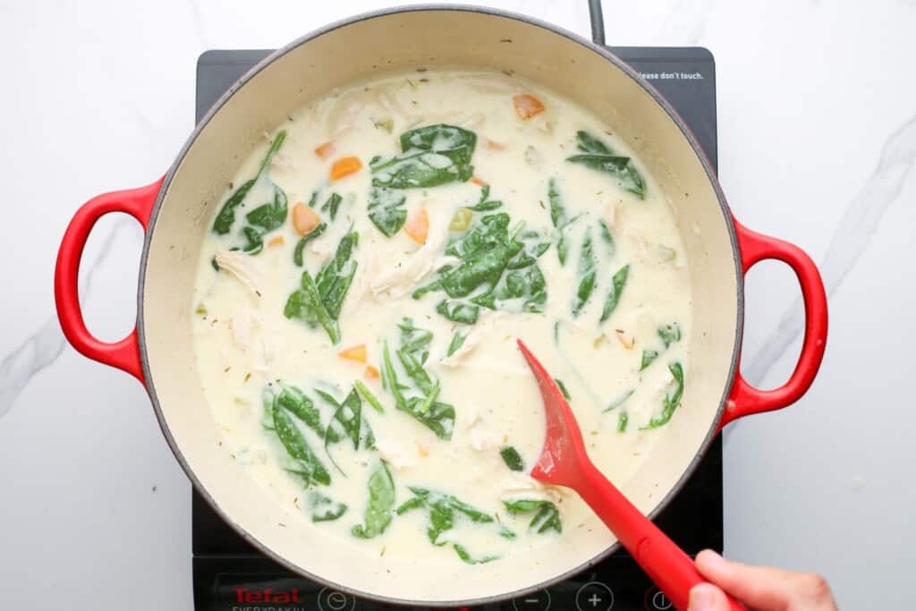 a dutch oven of creamy chicken soup with spinach stirred in with a red spoon.