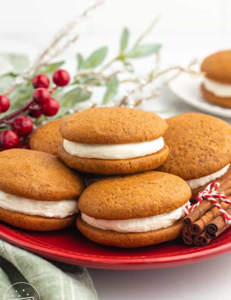 a red plate of gingerbread whoopie pies. A bundle of cinnamon decorates the plate and holiday greenery is in the background