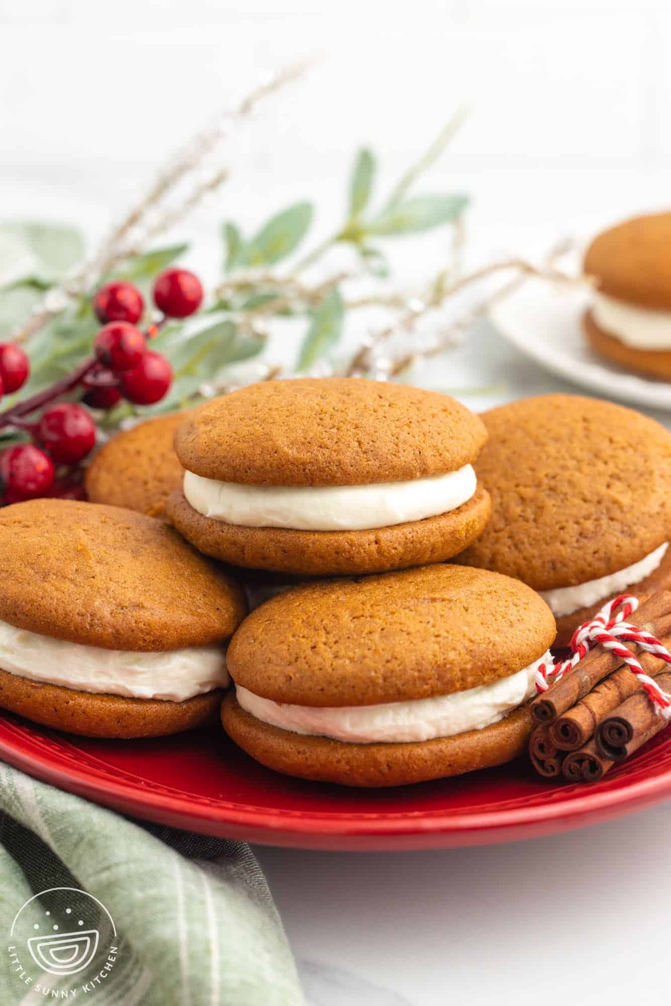 a red plate of gingerbread whoopie pies. A bundle of cinnamon decorates the plate and holiday greenery is in the background
