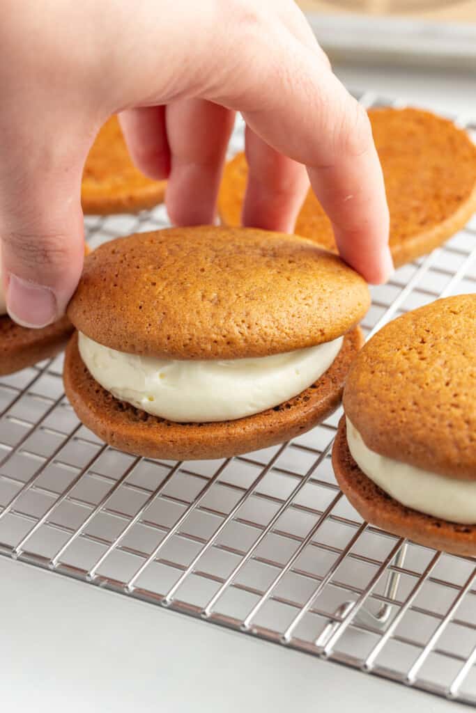 a hand adding the top piece to gingerbread whoopie pies on a wire rack.