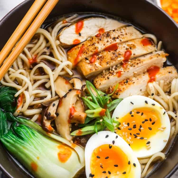 a black bowl of ramen soup with sliced chicken, soft boiled eggs, and veggies.