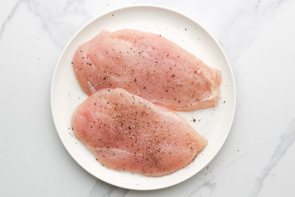 two raw chicken breast cutlets on a plate, seasoned with salt and pepper.