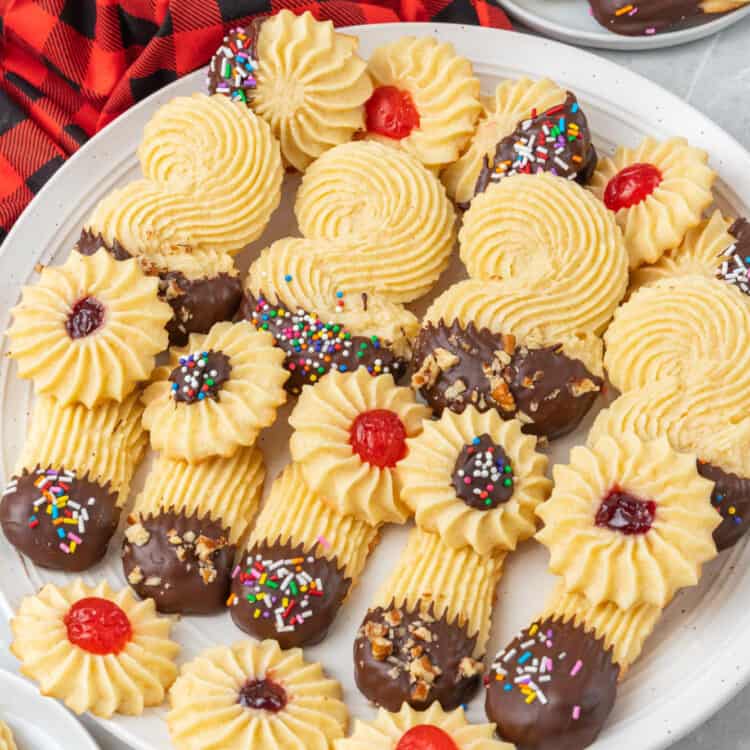 A white plate holding chocolate covered italian butter cookies in various textured shapes. Some cookies have sprinkles, come have candied cherries in the center.