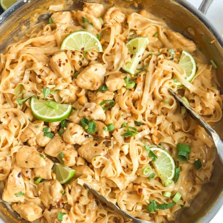 a skillet holding rice noodles with peanut sauce, chicken, and limes.