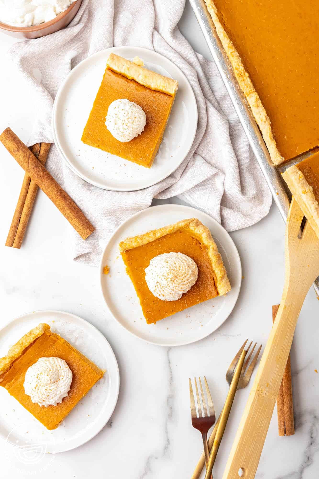 Overhead shot of small plates of square pieces of pumpkin slab pie topped with whipped cream, and sticks of cinnamon