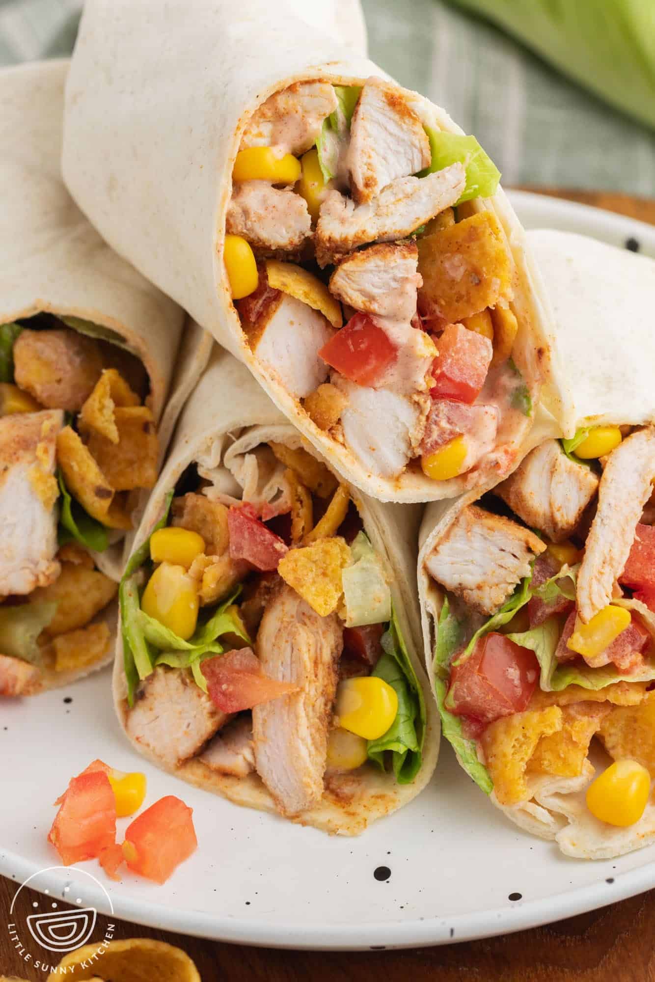 two chicken wraps, cut in half, on a plate. Inside of the wrap sandwiches is chicken, corn, tomatoes, lettuce, cheese, and fritos corn chips.