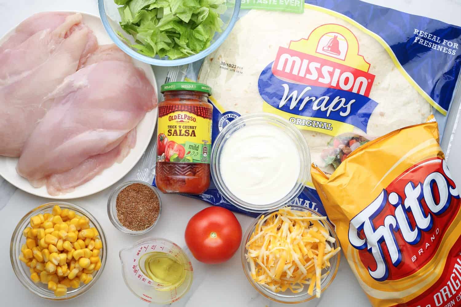 The ingredients needed to make southwest chicken wraps, with large tortillas, chicken breast, and toppings. A bag of fritos is also with the ingredients.